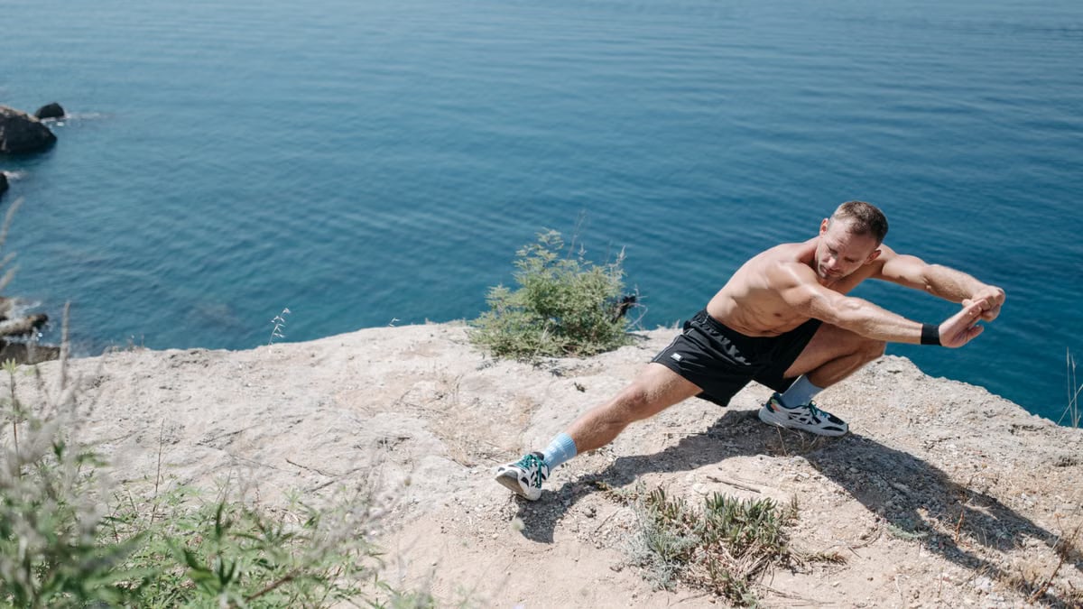 Shirtless man doing a stretch on a cliff overlooking the ocean in daylight.